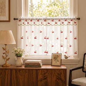 May include: A white cafe curtain featuring a repeating pattern of small red embroidered cherries. The curtain has a scalloped top edge with a thin green embroidered border and is hung on a dark brown rod across a window frame.