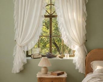 May include: A pair of white sheer curtains with ruffled edges hanging on a window. The curtains are tied back with simple cords, revealing a window frame with a view of green foliage. The fabric is lightweight and semi-transparent with ruffles.