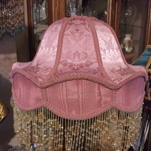 May include: A pink lampshade with a beaded fringe. The lampshade has a damask pattern and is trimmed with a pink ribbon.