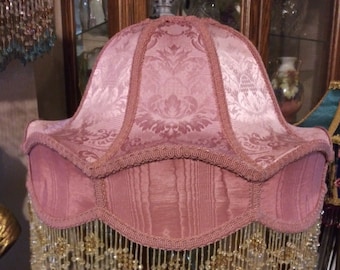 May include: A pink lampshade with a beaded fringe. The lampshade has a damask pattern and is trimmed with a pink ribbon.