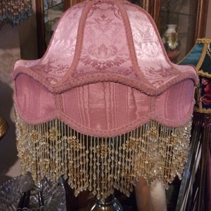 May include: A pink lampshade with a beaded fringe. The lampshade has a floral pattern and is made of fabric.
