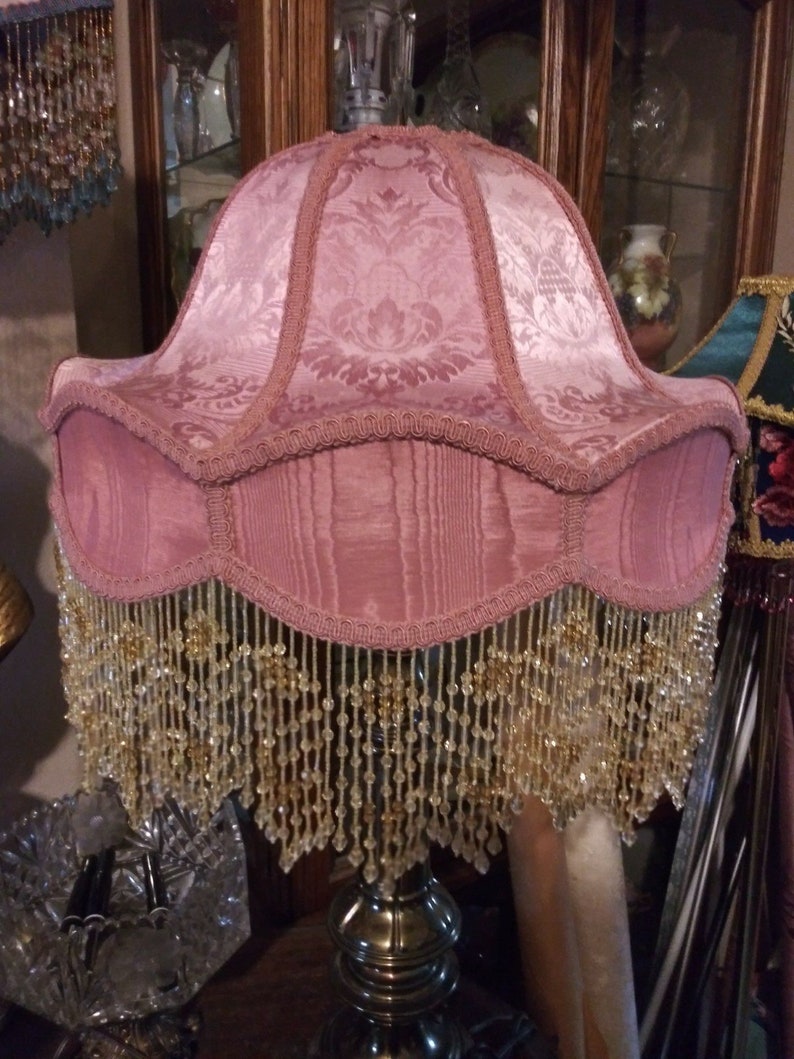 May include: A pink lampshade with a beaded fringe. The lampshade has a floral pattern and is made of fabric.