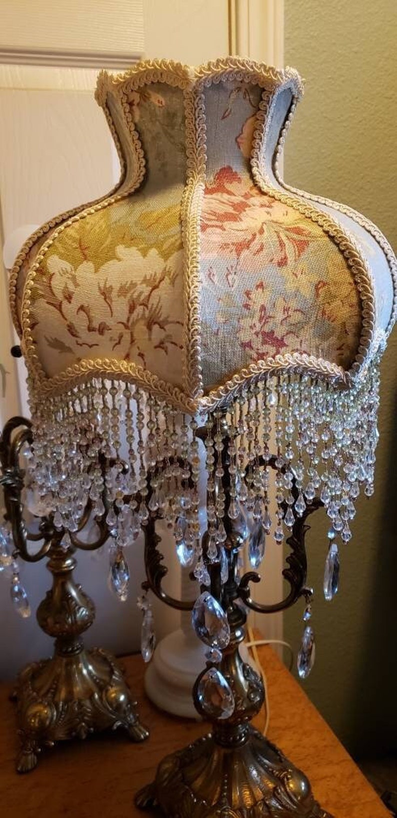 May include: A pair of ornate gold candle holders with crystal accents and a floral patterned lampshade with a beaded fringe.