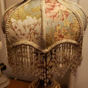 May include: A vintage table lamp with a floral patterned lampshade. The lampshade has a beaded fringe and a gold trim. The lamp base is made of brass and has a green and gold finish.
