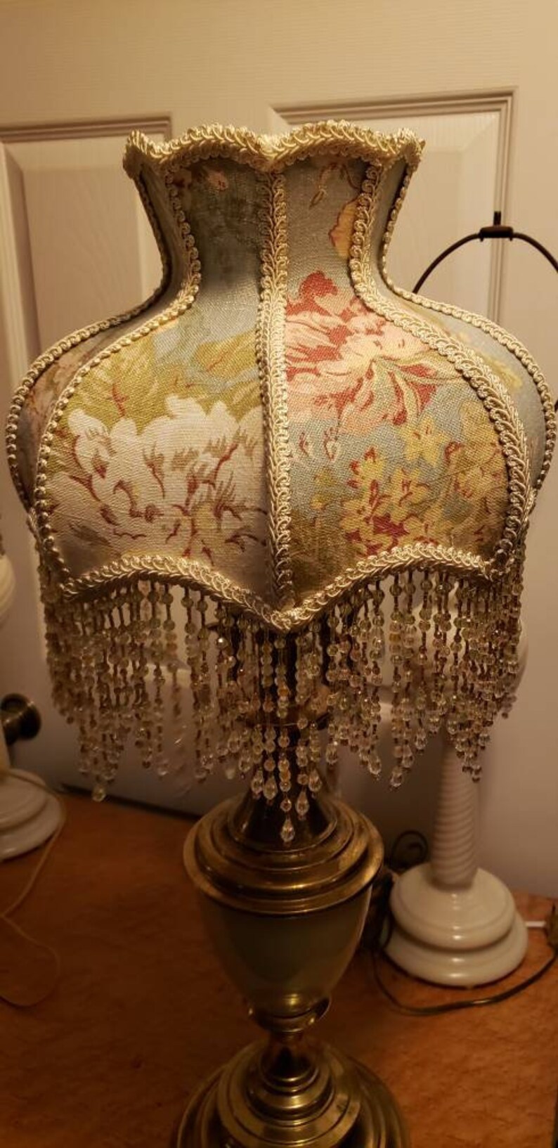 May include: A vintage table lamp with a floral patterned lampshade. The lampshade has a beaded fringe and a gold trim. The lamp base is made of brass and has a green and gold finish.