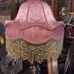 May include: A pink lampshade with a beaded fringe. The lampshade has a floral pattern and is made of fabric. The lampshade is on a silver lamp base.