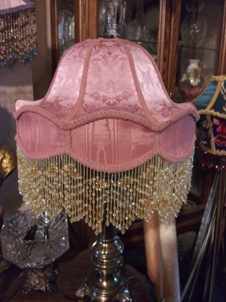 May include: A pink lampshade with a beaded fringe. The lampshade has a floral pattern and is made of fabric. The lampshade is on a silver lamp base.