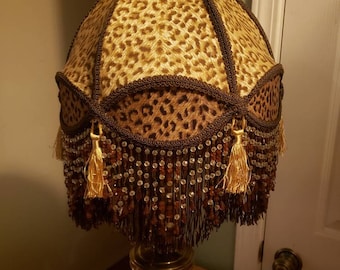 May include: A gold table lamp with a brown and beige leopard print lampshade with a beaded fringe and gold tassels.