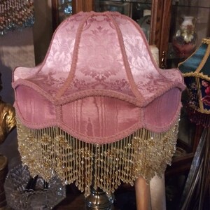 May include: A pink lampshade with a beaded fringe. The lampshade has a floral pattern and is made of fabric.
