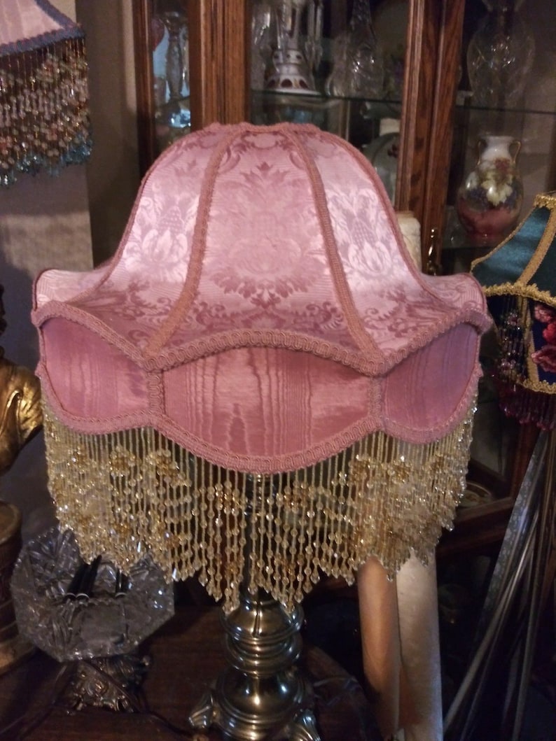 May include: A pink lampshade with a beaded fringe. The lampshade has a floral pattern and is made of fabric.
