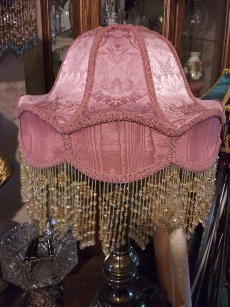 May include: A pink lampshade with a beaded fringe. The lampshade has a damask pattern and is trimmed with a pink ribbon.