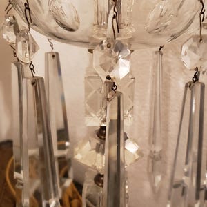 May include: Close-up of a crystal chandelier lamp. The lamp features a gold-colored base and a clear glass shade with hanging crystal prisms. The prisms are rectangular and diamond-shaped. The lamp is designed to be a decorative lighting fixture.
