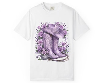 Puede incluir: Camiseta blanca con un diseño púrpura de un sombrero y botas de vaquero, rodeados de flores. Las botas y el sombrero tienen diseños detallados. Una cuerda está drapeada cerca de las botas, y también se incluye un pequeño jarrón de flores.