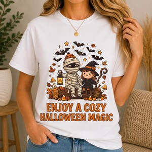 May include: A white short-sleeve t-shirt featuring a graphic of a mummy and a witch sitting on a log, surrounded by bats, stars, and autumn leaves. The text below the illustration reads, "ENJOY A COZY HALLOWEEN MAGIC" in bold orange letters.