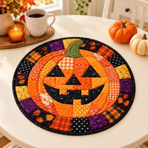 Peut inclure: Un tapis en tissu matelassé rond présentant un motif de citrouille d'Halloween en patchwork. La citrouille est composée de divers carrés de tissu à motifs orange, noirs et jaunes, avec une tige verte et un visage triangulaire noir.