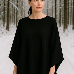 Op de afbeelding: Een zwarte poncho-achtige top met een ronde halslijn. De top heeft wijde mouwen en een losse pasvorm. Het model staat voor een besneeuwde bosachtergrond. De top is gemaakt van een zachte, soepele stof.