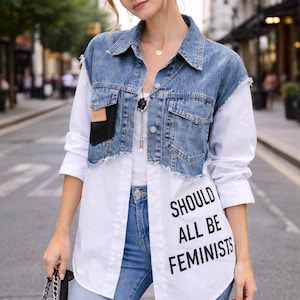Puede incluir: Chaqueta vaquera con dobladillo deshilachado sobre una camisa blanca. La camisa tiene el texto negro "SHOULD ALL BE FEMINISTS". La modelo lleva vaqueros azules y un bolso negro.
