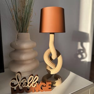 Könnte beinhalten: Dekorative Tischlampe mit kupferfarbenem Lampenschirm und Holzfuß mit Knotendesign. Eine Vase mit getrocknetem Gras und ein Holzschild mit der Aufschrift "Hall LOUNGE" befinden sich ebenfalls auf dem Tisch.