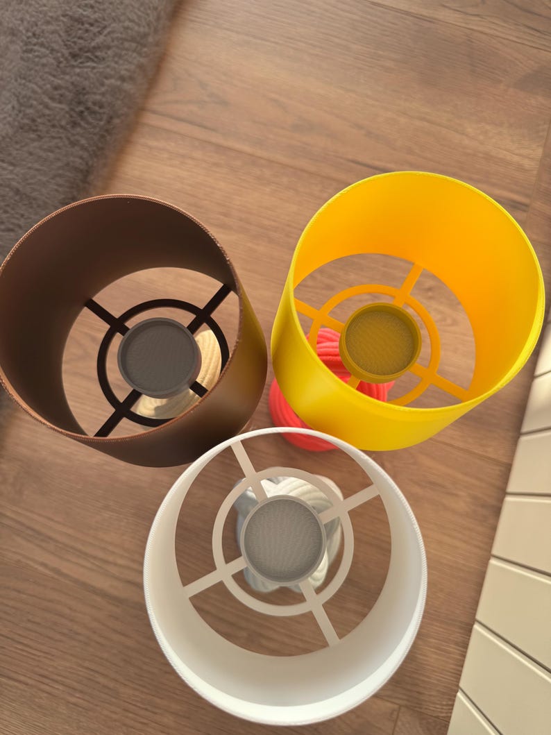 May include: Three colorful lampshades: a brown, a yellow, and a white one. The brown lampshade is cylindrical, the yellow one is a cone shape, and the white one is a cylinder. Each lampshade has a metal frame and a mesh screen at the bottom.