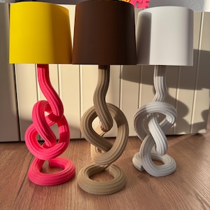 May include: Three decorative table lamps with unique, knotted bases. The lampshades are yellow, brown, and white. The bases are pink, beige, and white. The lamps are on a wooden surface.