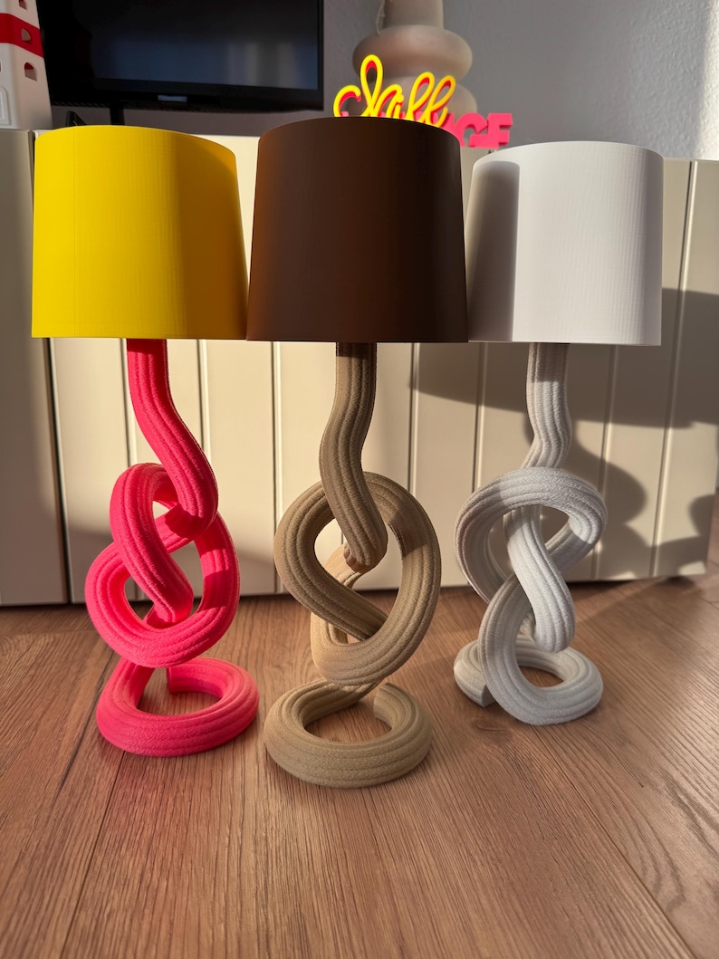 May include: Three decorative table lamps with unique, knotted bases. The lampshades are yellow, brown, and white. The bases are pink, beige, and white. The lamps are on a wooden surface.