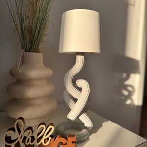 May include: A white table lamp with a cylindrical lampshade and a twisted, rope-like base. The lamp sits on a white surface next to a vase with dried plants and a wooden sign that reads "Shall Lounge."