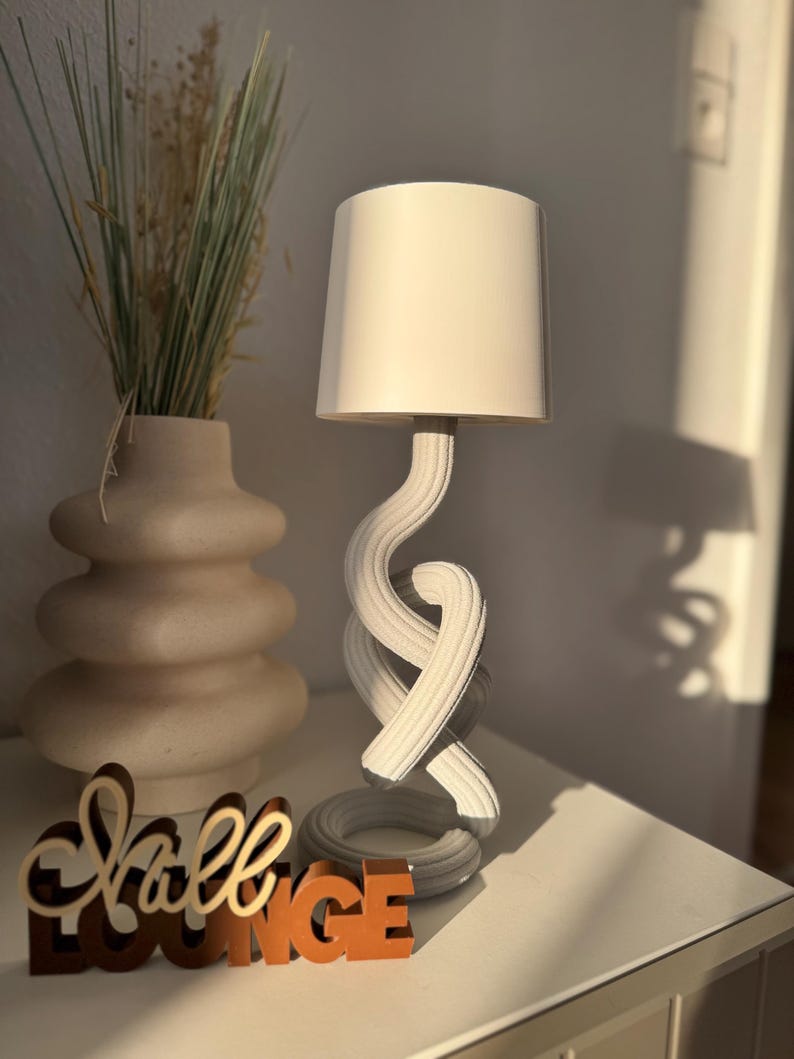 May include: A white table lamp with a cylindrical lampshade and a twisted, rope-like base. The lamp sits on a white surface next to a vase with dried plants and a wooden sign that reads "Shall Lounge."