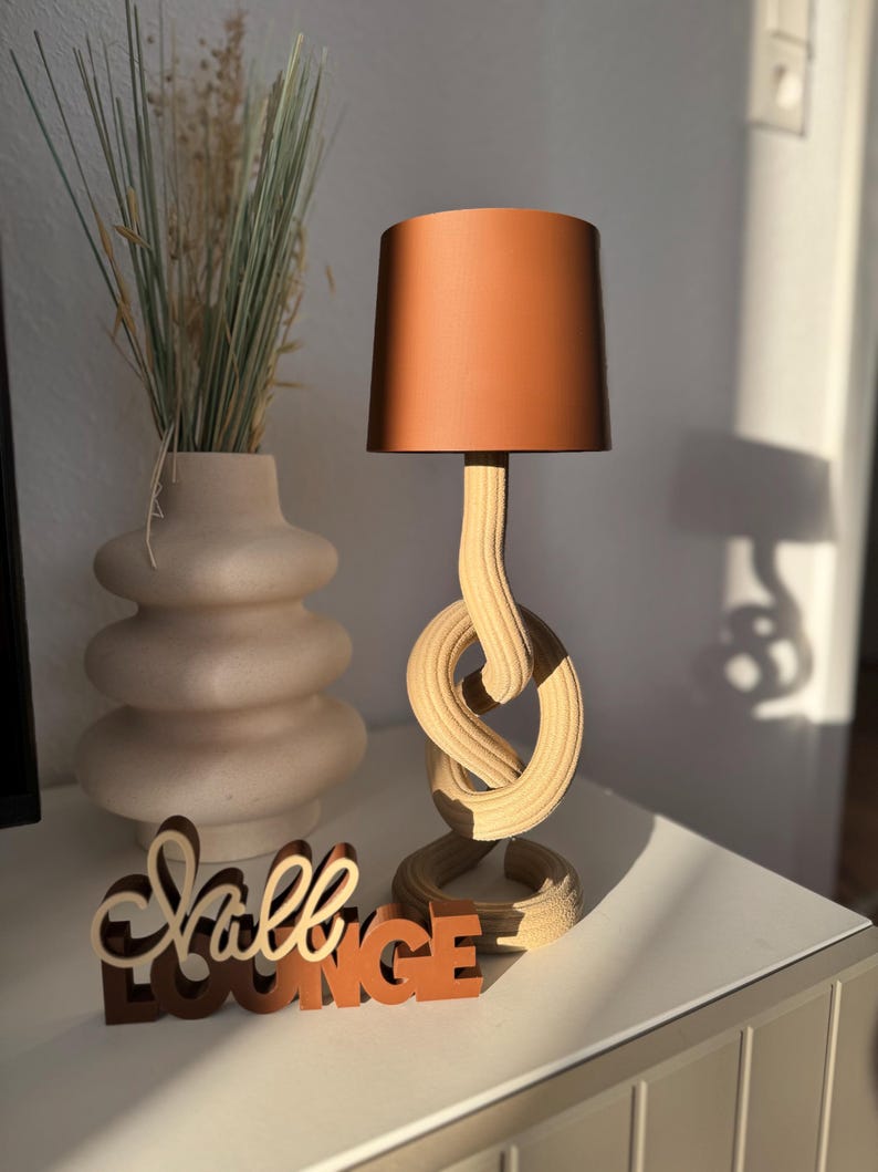 Könnte beinhalten: Dekorative Tischlampe mit kupferfarbenem Lampenschirm und Holzfuß mit Knotendesign. Eine Vase mit getrocknetem Gras und ein Holzschild mit der Aufschrift "Hall LOUNGE" befinden sich ebenfalls auf dem Tisch.