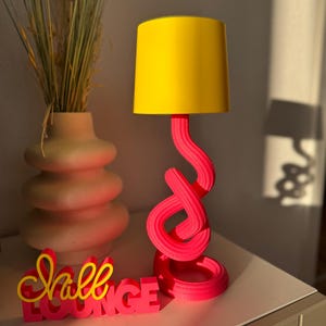 May include: A modern table lamp with a bright yellow lampshade and a twisted, hot pink base. The lamp sits next to a decorative vase with dried grass and a sign that reads "Chill Lounge" in pink and yellow.