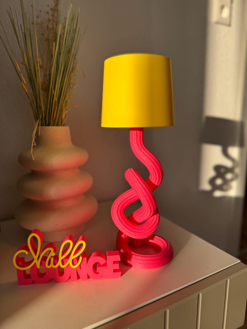 May include: A modern table lamp with a bright yellow lampshade and a twisted, hot pink base. The lamp sits next to a decorative vase with dried grass and a sign that reads "Chill Lounge" in pink and yellow.