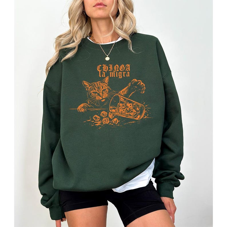 May include: Dark green sweatshirt featuring an orange graphic of a cat knocking over a glass of ice. The text "Chinga la migra" is above the cat illustration. The sweatshirt has long sleeves and a crew neck.