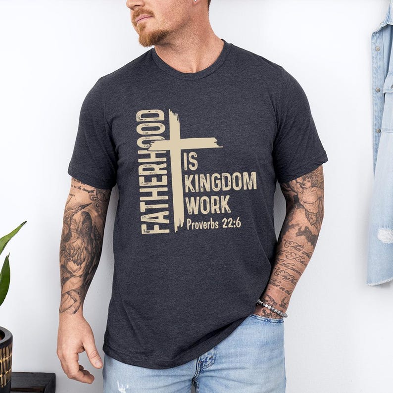 May include: Dark grey t-shirt featuring the text "FATHERHOOD IS KINGDOM WORK Proverbs 22:6" in a vertical and horizontal layout with a cross graphic. The shirt has short sleeves.