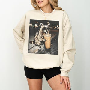 Peut inclure: Sweat-shirt cr&egrave;me avec un graphisme d'un raton laveur portant des lunettes de soleil et buvant un caf&eacute; glac&eacute;. Le raton laveur est assis sur un rebord. Le sweat-shirt a des manches longues et un col rond.