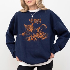 May include: Navy blue sweatshirt with an orange graphic of a cat knocking over a glass of ice. The text "CHINGA la migra" is above the cat. The sweatshirt is a crew neck style.