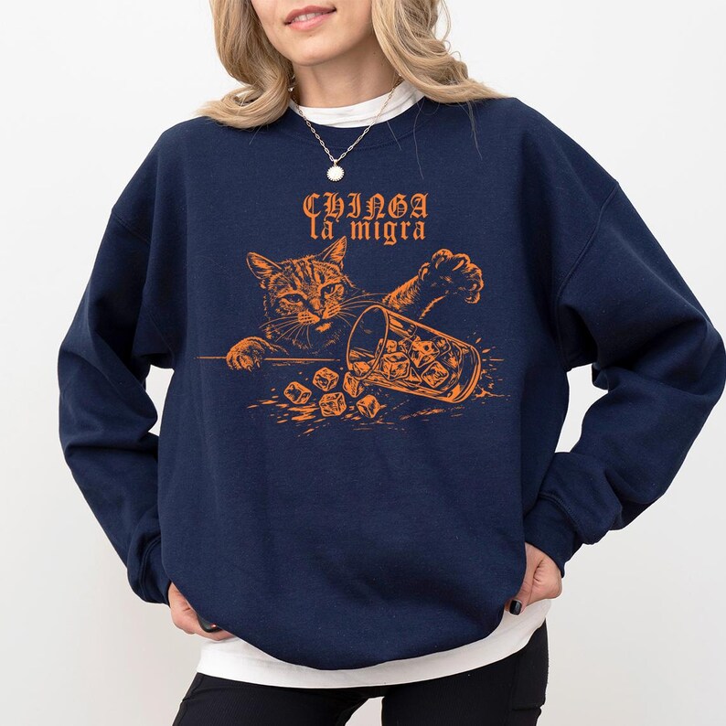 May include: Navy blue sweatshirt with an orange graphic of a cat knocking over a glass of ice. The text "CHINGA la migra" is above the cat. The sweatshirt is a crew neck style.