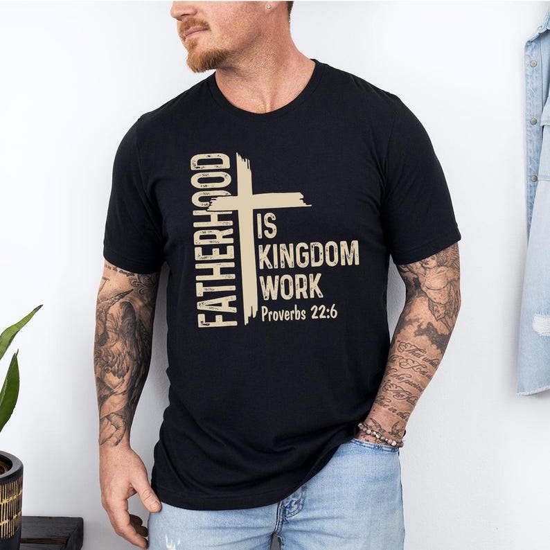May include: Black crew neck t-shirt featuring the text "FATHERHOOD IS KINGDOM WORK Proverbs 22:6" in beige. The text is arranged with a vertical element on the left and a cross graphic.