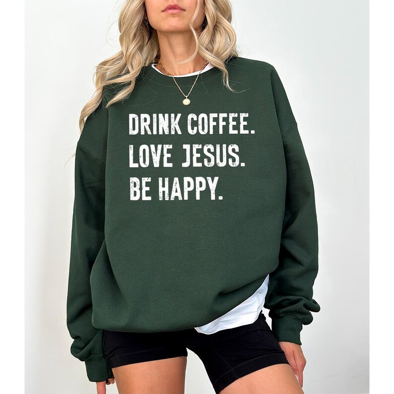 May include: Dark green sweatshirt with the text "DRINK COFFEE. LOVE JESUS. BE HAPPY." in white block letters. The sweatshirt has long sleeves and a crew neck. The model is wearing black shorts and a white undershirt.