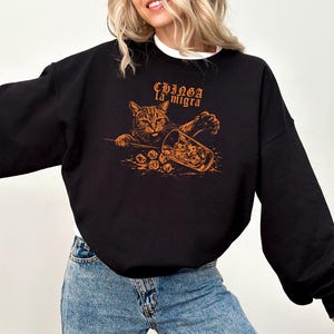 May include: Black sweatshirt with an orange graphic of a cat reaching for a glass of ice. The text "CHINGA la migra" is above the cat. The sweatshirt is worn by a person wearing blue jeans.