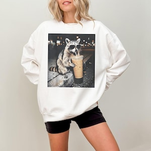Peut inclure: Sweat-shirt blanc avec un dessin de raton laveur portant des lunettes de soleil et buvant du caf&eacute; glac&eacute;. Le raton laveur est repr&eacute;sent&eacute; dans un cadre urbain nocturne. Le sweat-shirt est de style d&eacute;contract&eacute;.