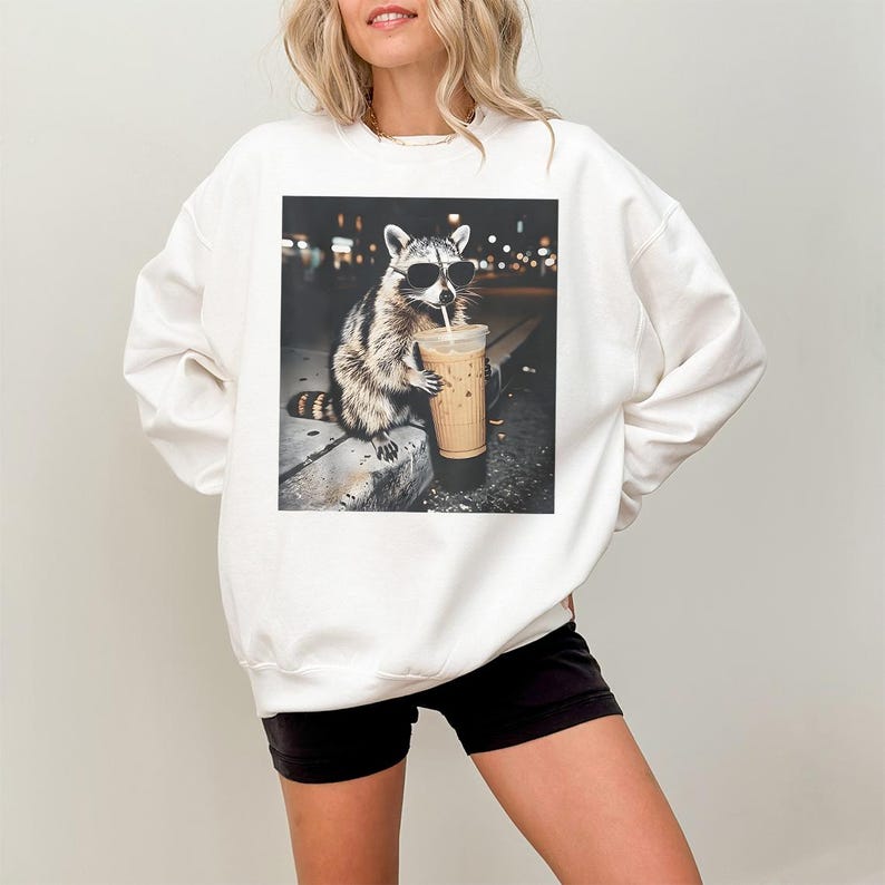 Peut inclure: Sweat-shirt blanc avec un dessin de raton laveur portant des lunettes de soleil et buvant du caf&eacute; glac&eacute;. Le raton laveur est repr&eacute;sent&eacute; dans un cadre urbain nocturne. Le sweat-shirt est de style d&eacute;contract&eacute;.