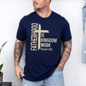 May include: Navy blue t-shirt with the text "FATHERHOOD IS KINGDOM WORK Proverbs 22:6" in a beige colour. The design includes a cross. The shirt is short-sleeved.