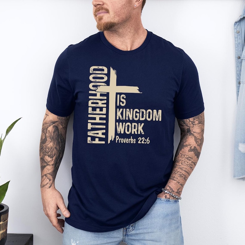 May include: Navy blue t-shirt with the text "FATHERHOOD IS KINGDOM WORK Proverbs 22:6" in a beige colour. The design includes a cross. The shirt is short-sleeved.