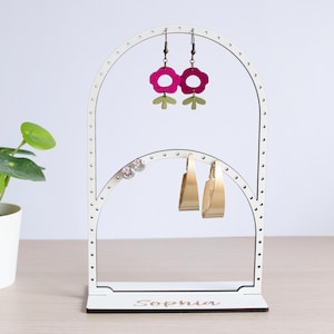 May include: A white earring display stand with a floral design, holding pink and gold earrings. A small potted plant is to the left. The stand has the name "Sophia" on the base.