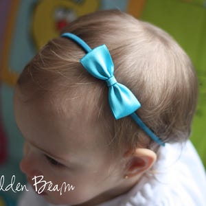May include: A thin, bright turquoise headband featuring a small, centered satin bow in the same vibrant color. The accessory is positioned across the top of the head, showcasing a smooth, shiny finish on the bow and a matching fabric-covered band.