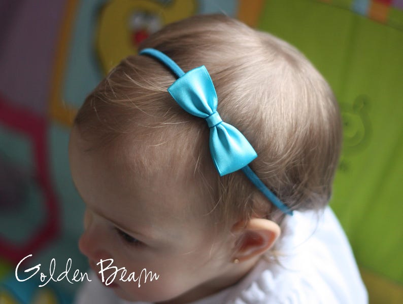 May include: A thin, bright turquoise headband featuring a small, centered satin bow in the same vibrant color. The accessory is positioned across the top of the head, showcasing a smooth, shiny finish on the bow and a matching fabric-covered band.