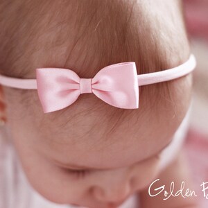 May include: A light pink satin bow headband featuring a smooth, shiny finish on the bow and a thin, matching elastic band. The text Golden Beam is visible in the bottom right corner of the image, written in a white, cursive script font.