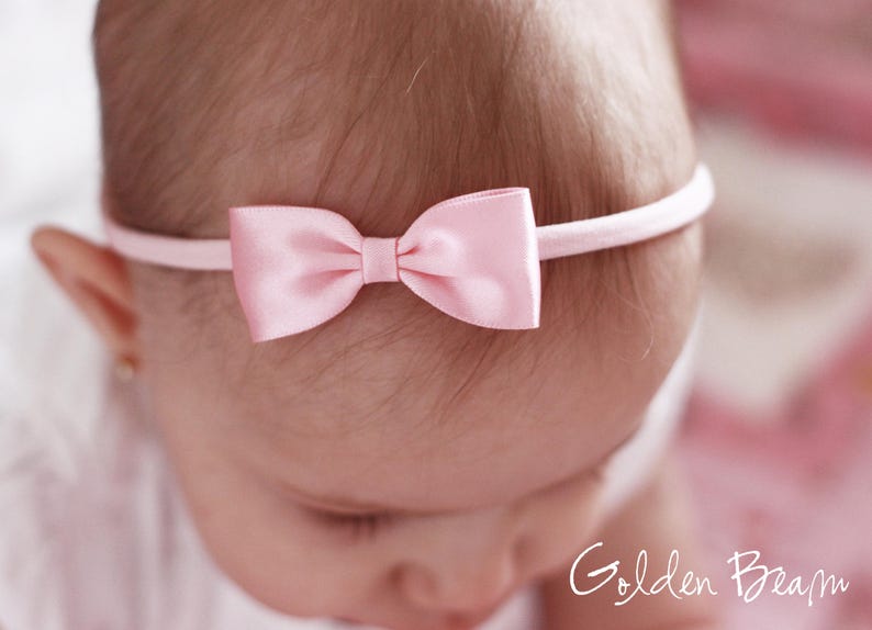May include: A light pink satin bow headband featuring a smooth, shiny finish on the bow and a thin, matching elastic band. The text Golden Beam is visible in the bottom right corner of the image, written in a white, cursive script font.