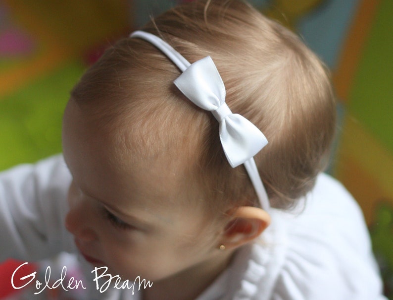 May include: A white satin bow headband for babies and toddlers. The headband is made of a soft, stretchy material and is perfect for everyday wear.
