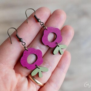 May include: A pair of handmade earrings featuring a flower design. The earrings have a magenta flower shape with a green leaf accent. The metal hardware is a dark bronze color. The earrings are lightweight and suitable for everyday wear.