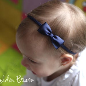May include: A thin, dark navy blue elastic headband featuring a small, centered satin bow. The accessory is positioned across the top of the head, showcasing a smooth, reflective fabric texture on the bow and a simple, minimalist band design.